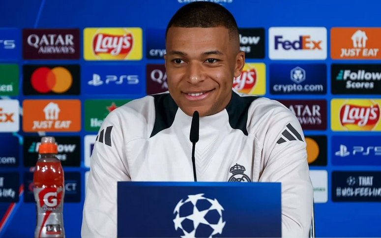 Kylian Mbappé sonriente en una conferencia de prensa, vistiendo una camiseta del Real Madrid, con un fondo de logos de patrocinadores y la insignia de la Liga de Campeones.