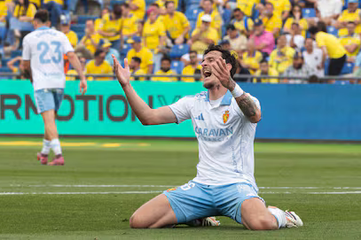 Jugador de fútbol arrodillado en el campo, con una expresión de frustración, mientras el público lleva ropa amarilla de fondo.