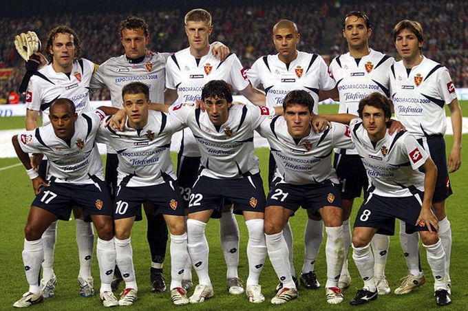 Equipo de fútbol posando en formación, con una mezcla de jugadores de pie y agachados, usando camisetas blancas y pantalones oscuros.