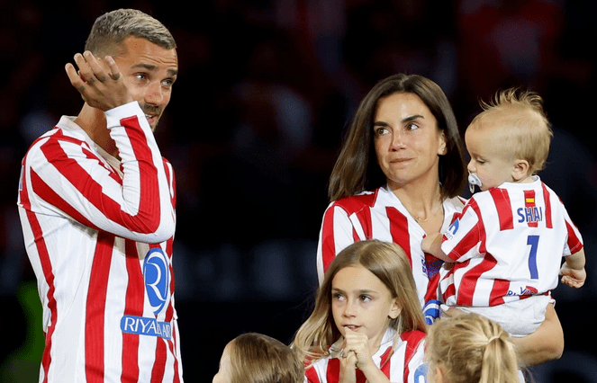 Un jugador de fútbol de pie junto a su familia, todos vestidos con camisetas de rayas rojas y blancas. El jugador hace un gesto mientras su pareja sostiene a un bebé. Hay dos niñas cerca, una de ellas mirando a la cámara.