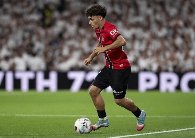 Un jugador de fútbol en una cancha, vistiendo una camiseta roja con negro, en pleno movimiento mientras controla un balón.